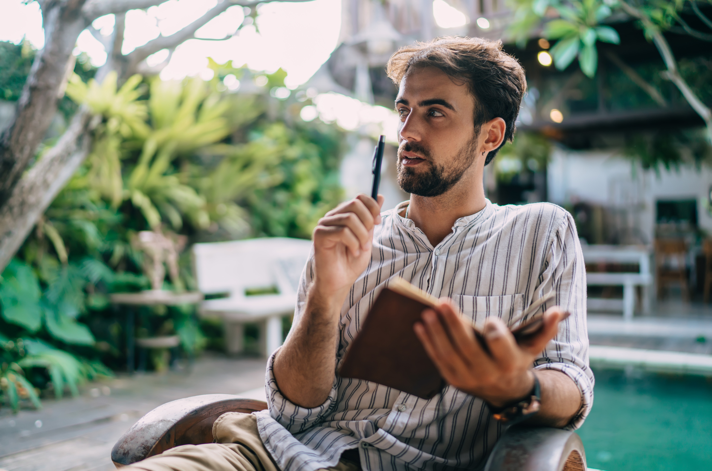 Wistful male author sitting with pen and notebook on poolside